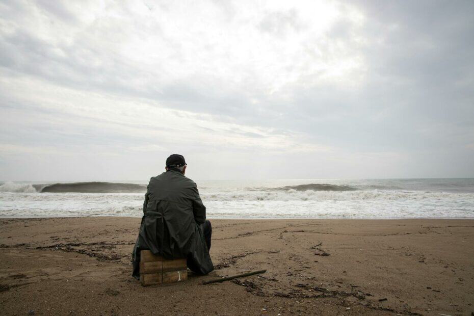 A man sitting alone on an empty beach looking at the stormy sea, evoking thoughts of solitude and reflection.