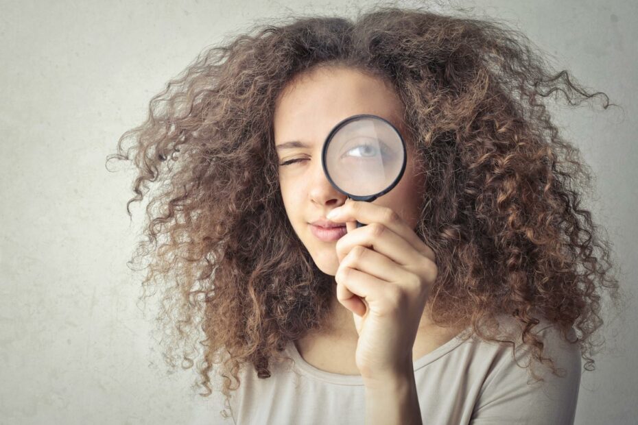 Close-up portrait of a woman winking and holding a magnifying glass against a white background.