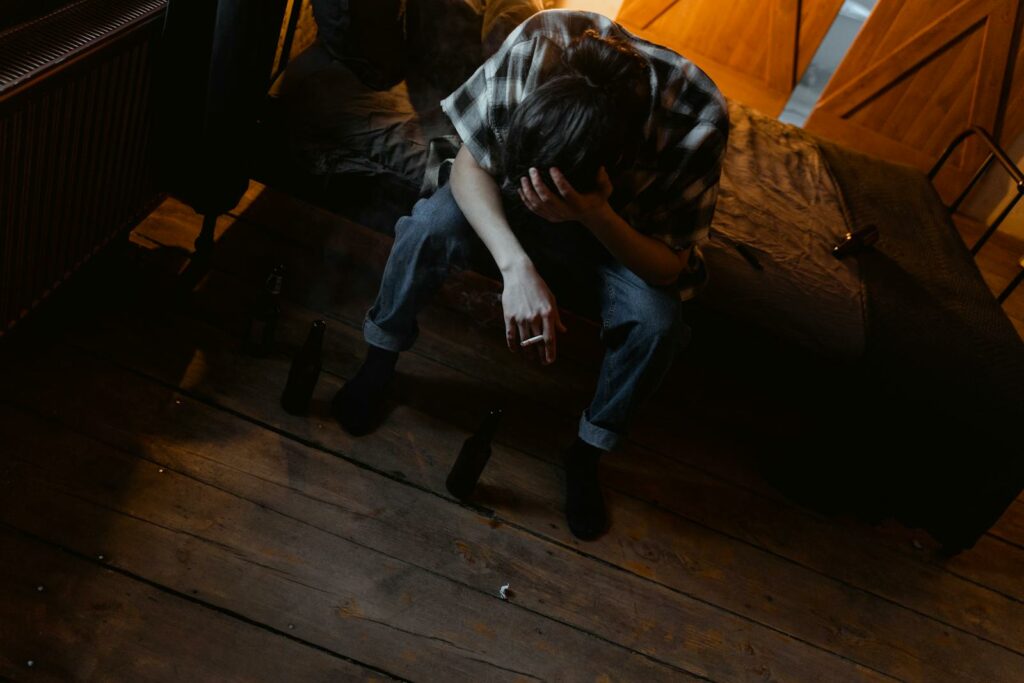 A young man sitting on a bed appears distressed, holding a cigarette in a dimly lit room.
