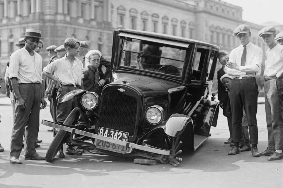 A vintage car crash attended by a group of onlookers in a 1920s urban setting.