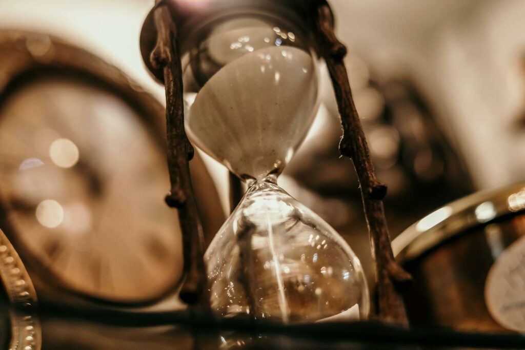 Close-up of a vintage hourglass with blurred antique clock background, evoking a timeless feel.