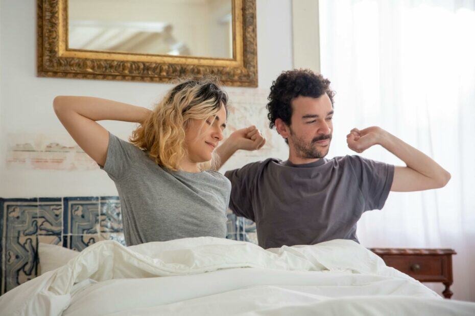 A couple begins their morning stretching in bed with soft daylight filtering through curtains.