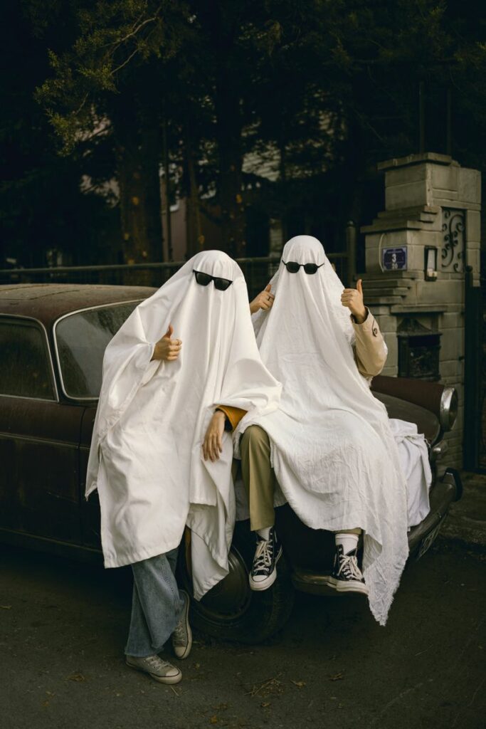 Three people dressed as ghosts with sunglasses, sitting on a vintage car, showing thumbs up.