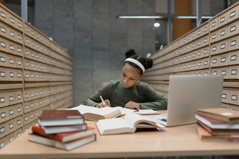 Dedicated African American girl studying in a library with books and a laptop.
